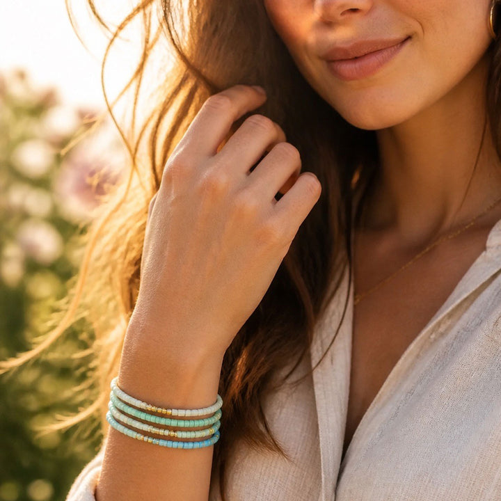 A close-up lifestyle shot of a woman outdoors wearing a stack of four beaded bracelets on her wrist. The bracelets feature light blue, mint green, and metallic gold beads.