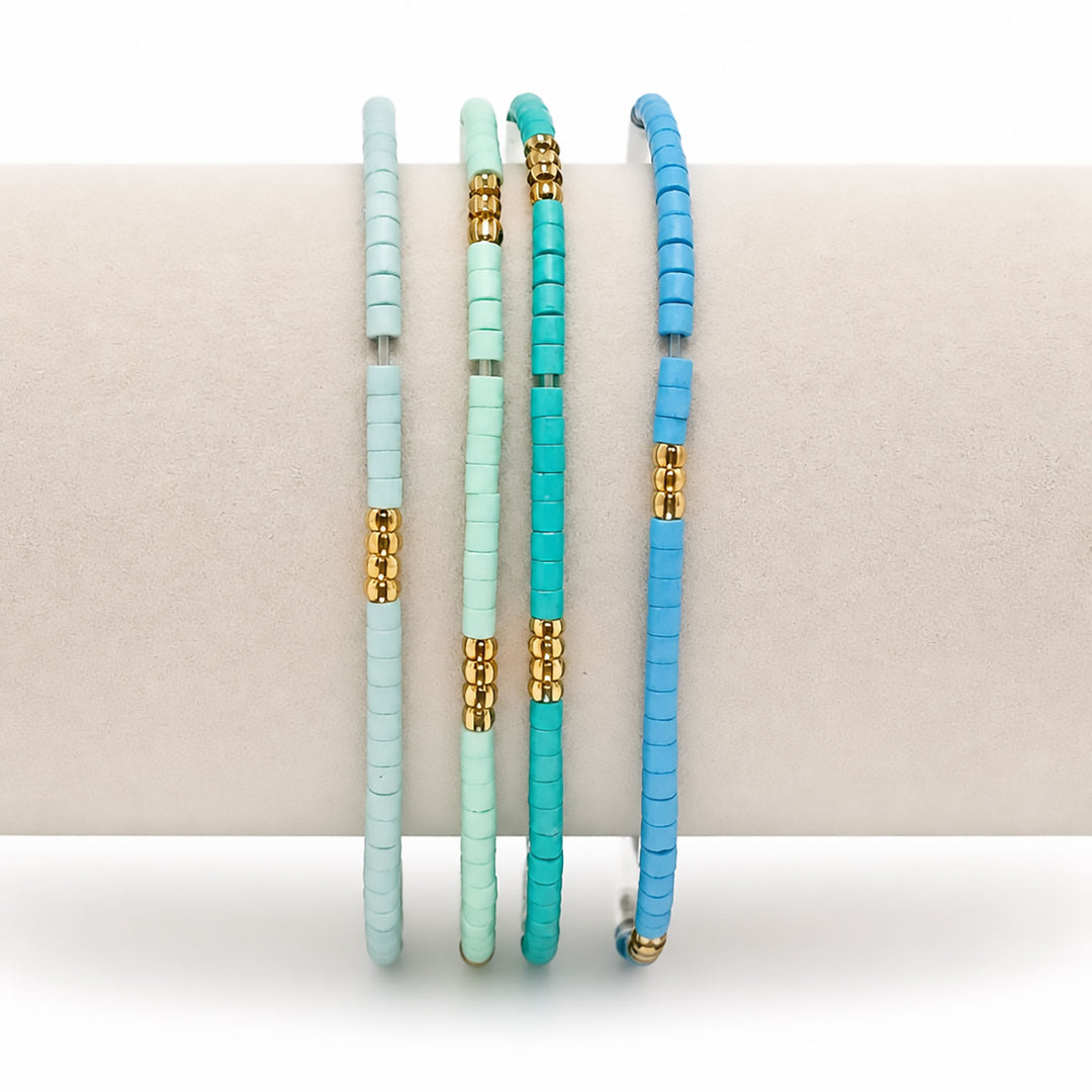 Three beaded bangles in pastel mint, turquoise, and bright blue, each accented with gold spacer beads, displayed on a neutral cream-colored stand.