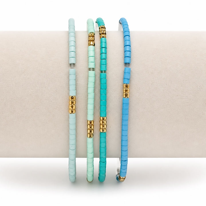 Three beaded bangles in pastel mint, turquoise, and bright blue, each accented with gold spacer beads, displayed on a neutral cream-colored stand.