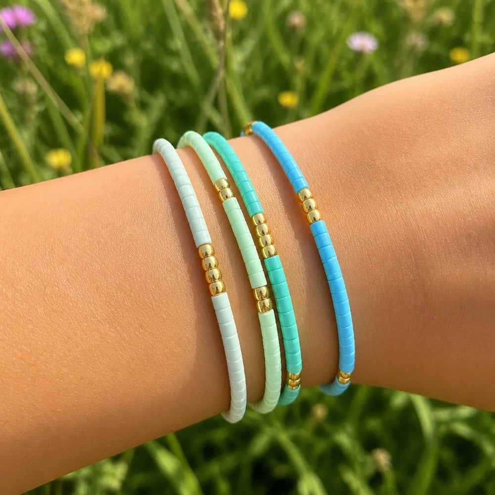 Four beaded stretch bracelets in pastel white, mint green, light blue, and sky blue, accented with gold spacer beads, shown worn on a wrist against a blurred green grassy background.