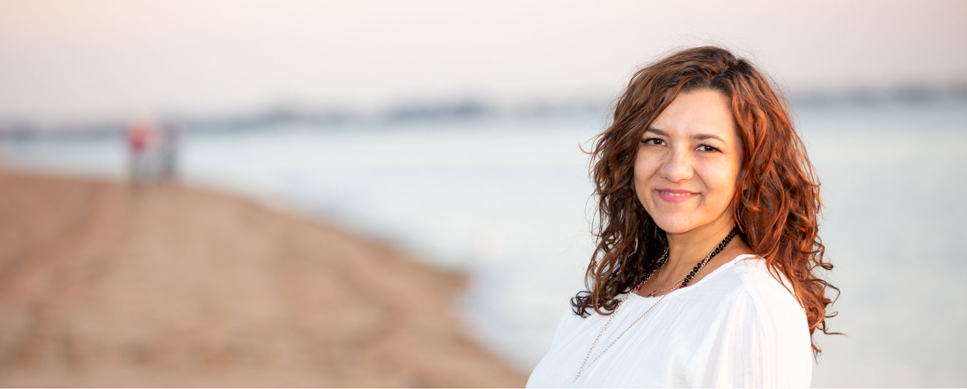 A woman with wavy brown hair wearing a white top, standing on a sandy beach with a calm body of water and a soft sunset sky in the background.