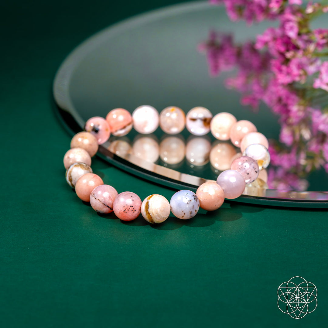 Multicolored beaded bracelet on a green surface with a blurred pink flower arrangement in the background.