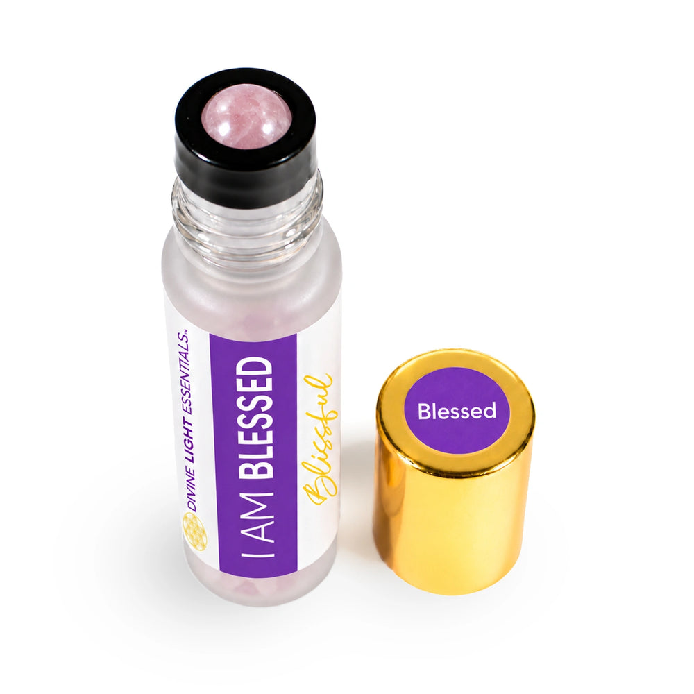 A clear glass roll-on bottle with a black cap and a pink rose quartz roller ball, shown next to its gold-colored metal cap. The bottle features a white and purple label that reads 'I AM BLESSED' and 'Divine Light Essentials'.