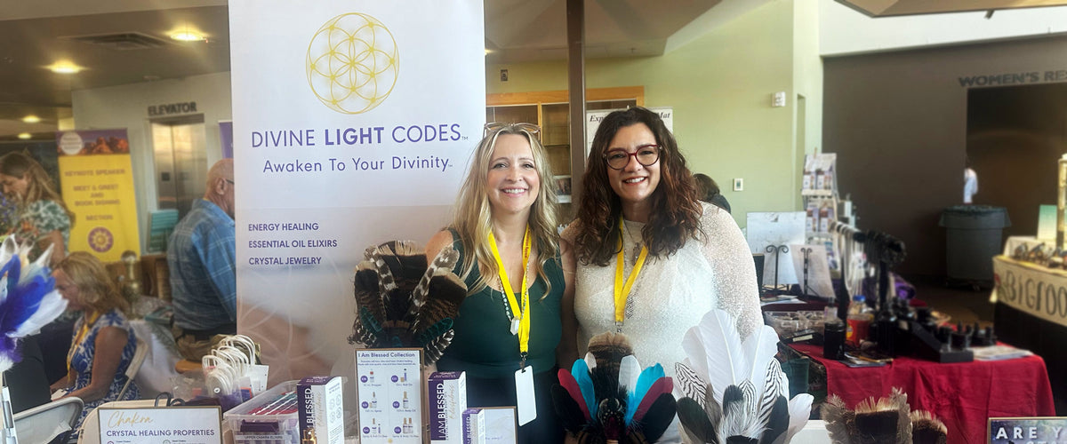 Two women smiling behind a display table at an event, with a white banner reading 'DIVINE LIGHT CODES. Awaken To Your Divinity.