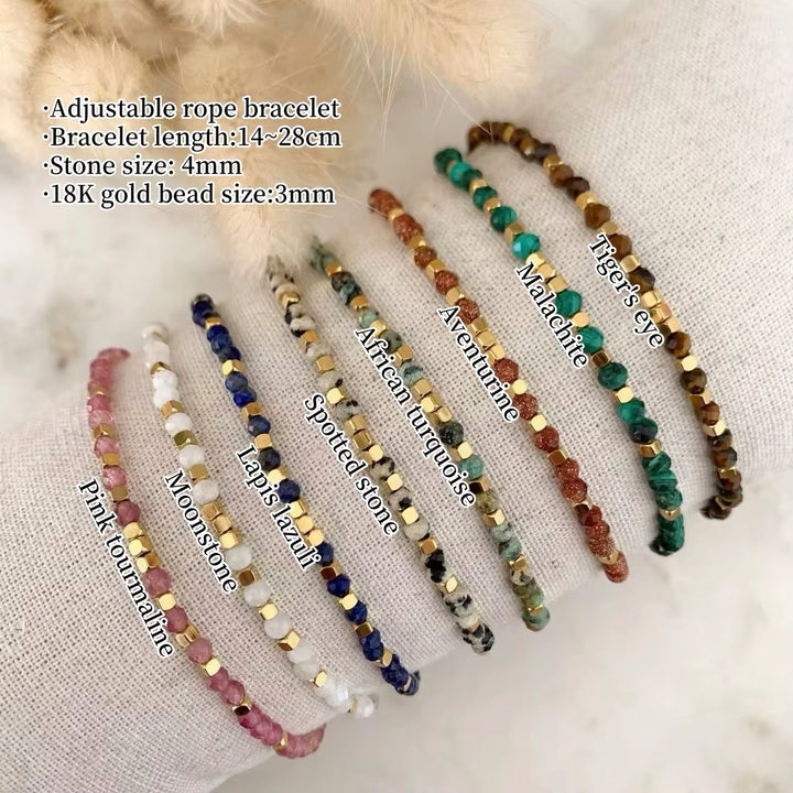 Eight adjustable rope bracelets made of faceted gemstone beads and gold spacer beads are arranged on a textured beige fabric background. The bracelets feature various stone colors including pink, white, navy, green, brown, and gold.