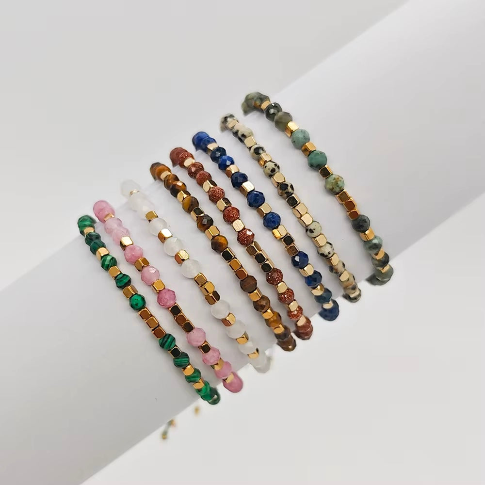 A set of eight beaded stretch bracelets arranged on a white surface. The bracelets feature various gemstone beads in green, pink, brown, blue, white, and gold-toned spacer beads.