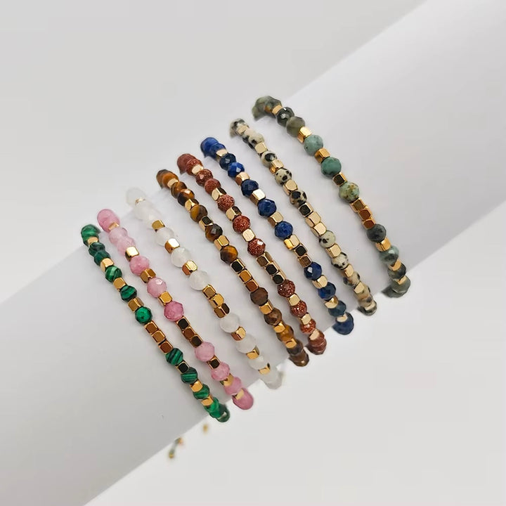 A set of eight beaded stretch bracelets arranged on a white surface. The bracelets feature various gemstone beads in green, pink, brown, blue, white, and gold-toned spacer beads.