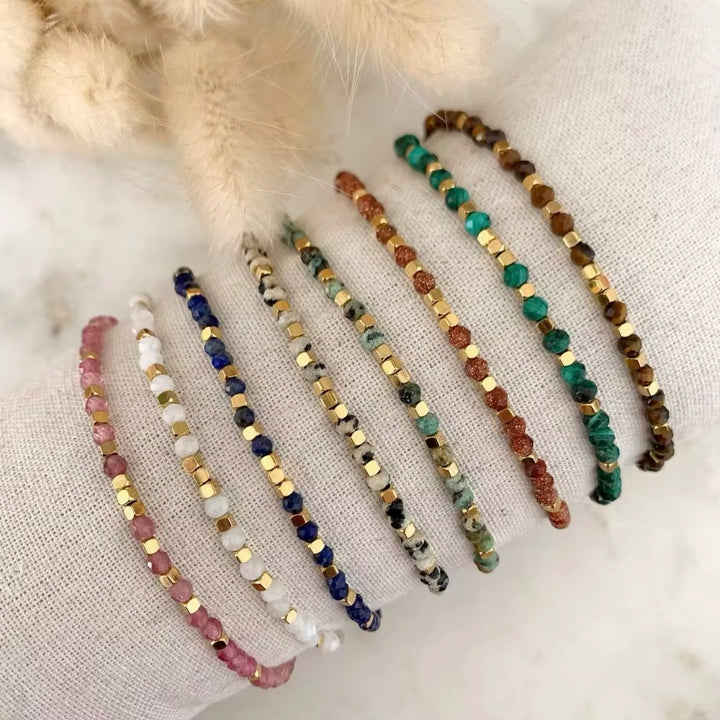 Six beaded stretch bracelets displayed on a textured white fabric surface, with a soft beige faux-fur prop at the top. The bracelets feature various gemstone beads in pink, white, blue, gray, green, and brown, accented with gold-toned spacer beads.