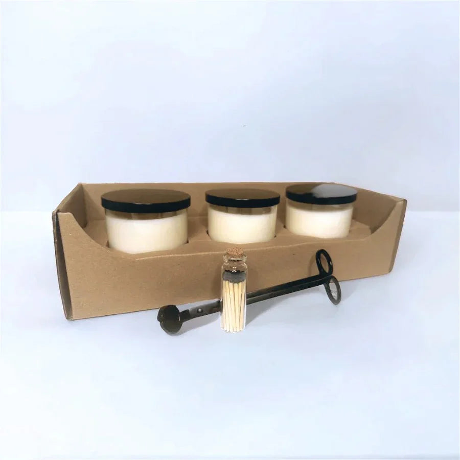 Three candles in a cardboard holder with a candle wick trimmer on a light blue background