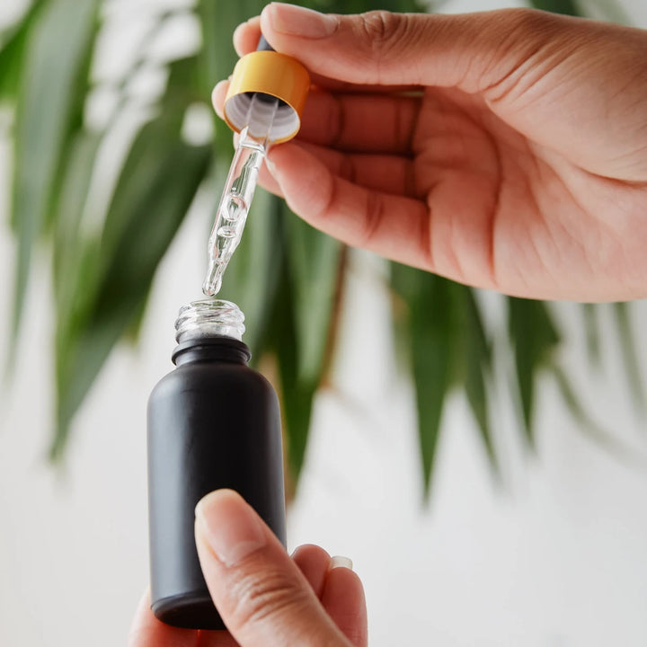 Hand holding dropper with clear liquid over black bottle, green plant background for healing wellness