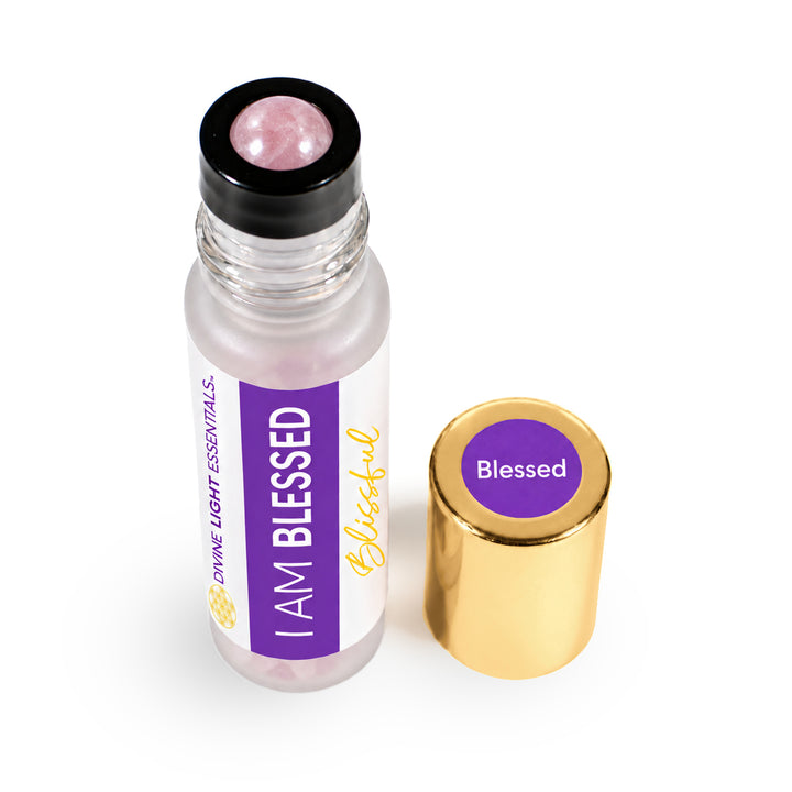 A clear glass roll-on bottle with a black cap and a pink rose quartz roller ball, shown next to its gold-colored metal cap. The bottle features a white and purple label that reads &