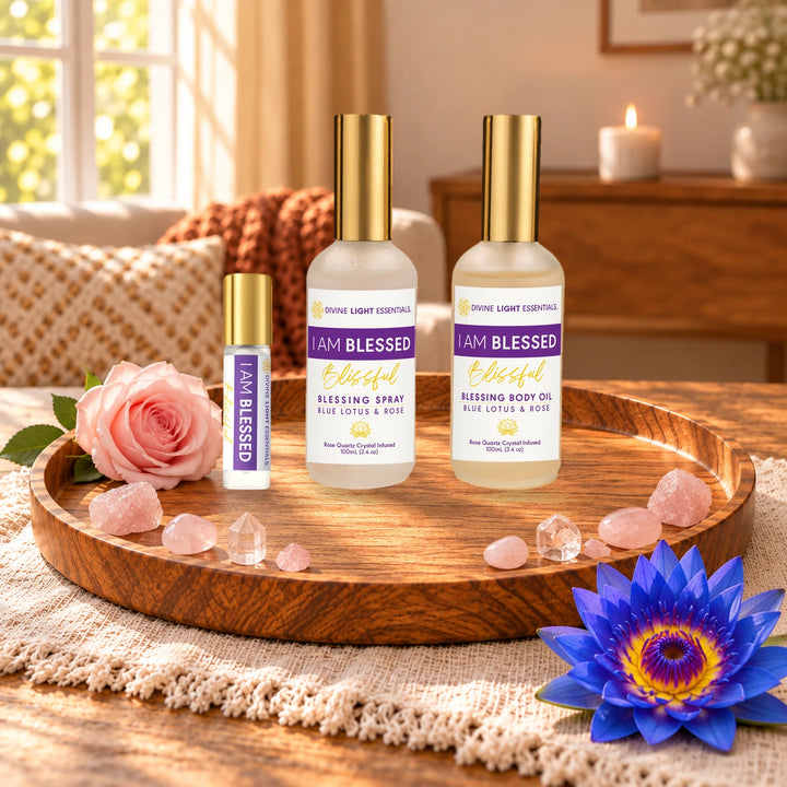 Two bottles of I Am Blessed Blissful essential products, including a small roller bottle and two larger spray/body oil bottles, arranged on a wooden tray with pink roses, crystals, and a blue lotus flower in a warm, softly lit room.