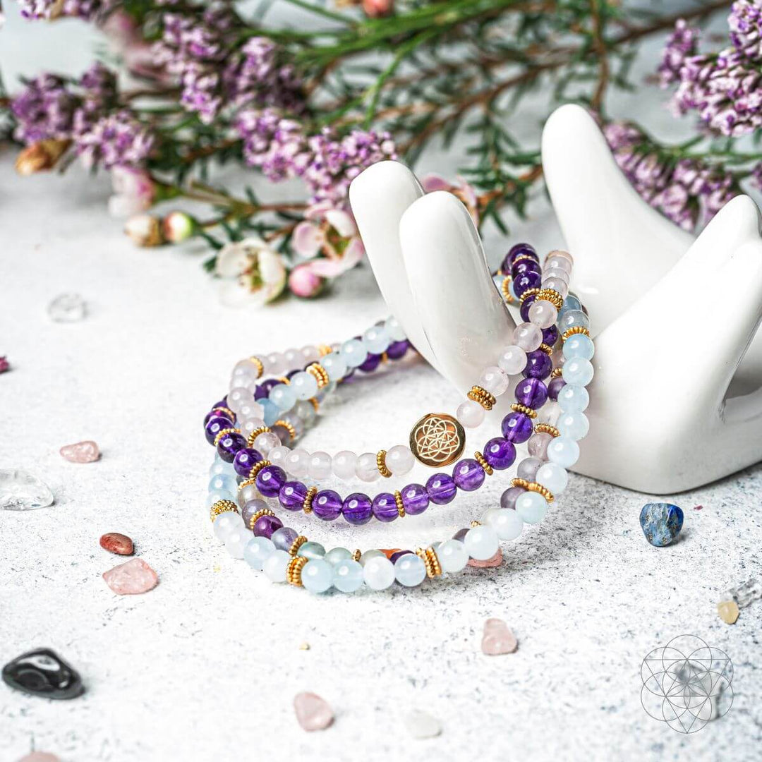 Stack of beaded bracelets with purple and gold accents on a white surface with flowers in the background