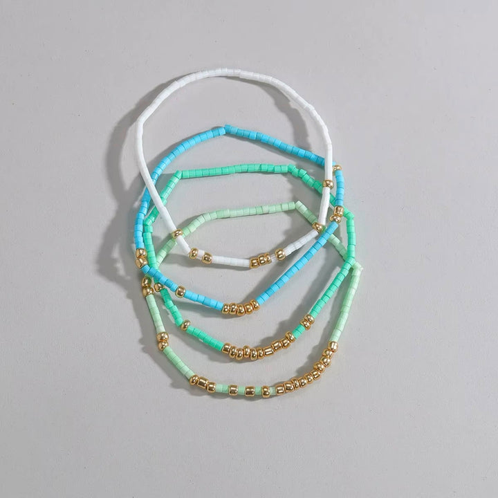 A set of five beaded stretch necklaces in white, light blue, mint green, and light purple, accented with small gold spacer beads, arranged on a neutral gray background.